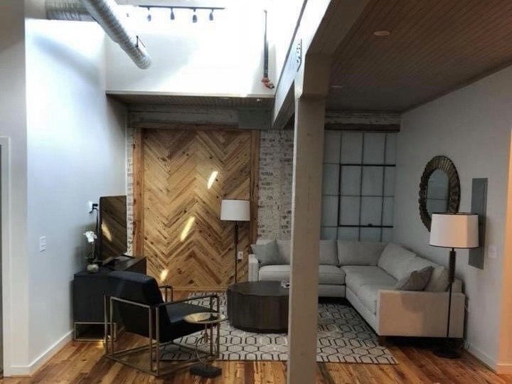 Open living area with a herringbone wood accent wall, exposed brick, a light gray sectional sofa, two floor lamps, track lighting and wide-plank wood floors