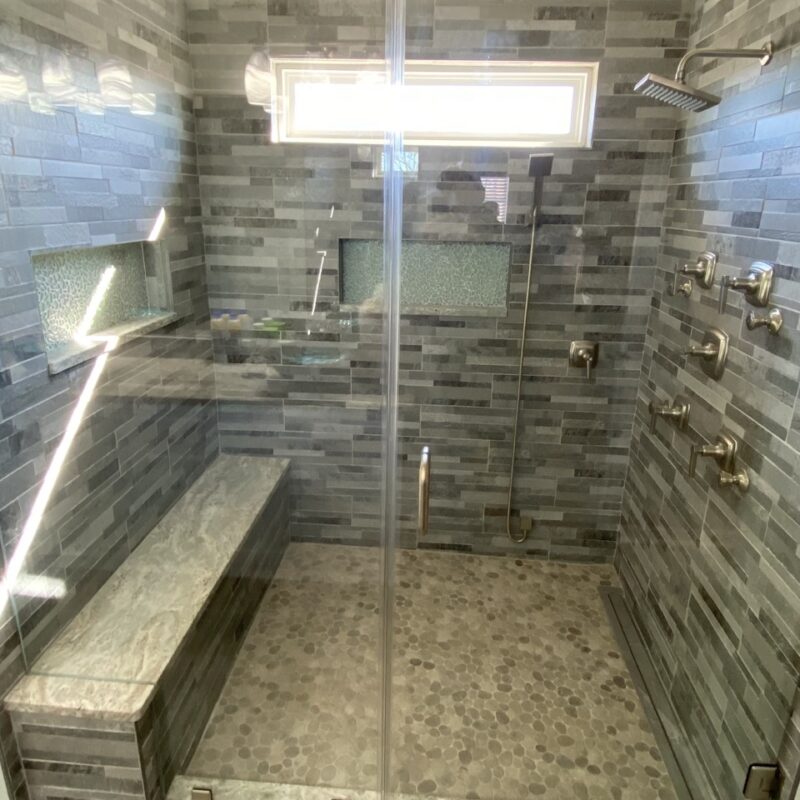 Luxury walk-in shower with glass enclosure, grey tile walls, built-in bench, and multiple shower heads.