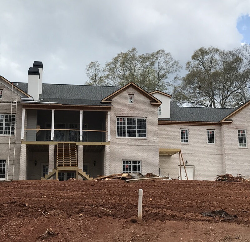 Two-story brick and wood home under construction with large covered porch and exposed soil.